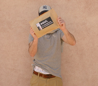 Hombre vestido con una camiseta gris y pantalones color caqui, usando una gorra de béisbol, sostiene un sobre de papel marrón que cubre su rostro. El sobre tiene etiquetas que incluyen texto promocional y logos