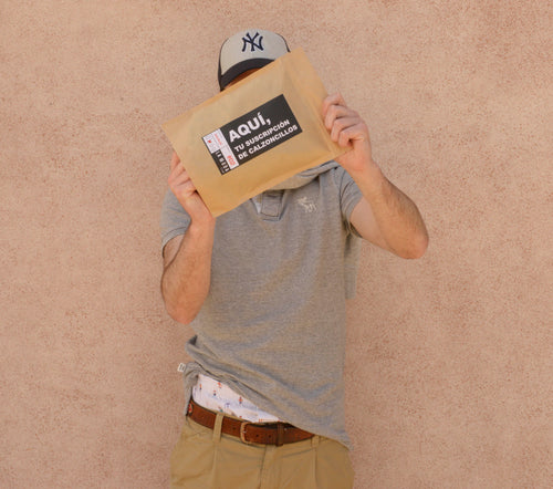 Hombre vestido con una camiseta gris y pantalones color caqui, usando una gorra de béisbol, sostiene un sobre de papel marrón que cubre su rostro. El sobre tiene etiquetas que incluyen texto promocional y logos