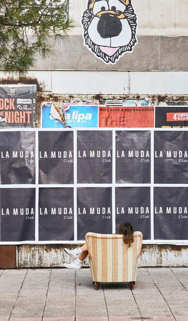 Portada movil para la muda. Una mujer sentada en un sillon observando un mural de pancartas de la muda club