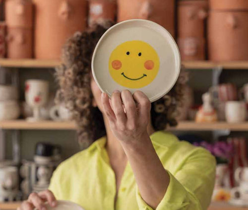 Una mujer sujetando un plato hecho en cerámica artesanal con el dibujo de una cara feliz estilo emoticono