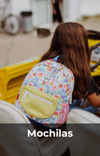 Niña con cabello largo sentada en un coche amarillo lleva una mochila estampada con flores y elementos coloridos, con un bolsillo frontal amarillo claro. Texto 'Mochilas' al pie de la imagen.