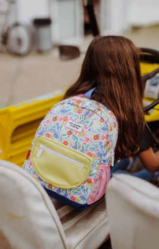 Niña con cabello largo sentada en un coche amarillo lleva una mochila estampada con flores y elementos coloridos, con un bolsillo frontal amarillo claro