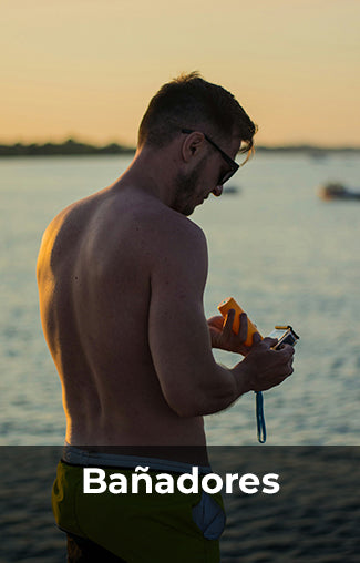 Hombre con gafas de sol y bañador verde aplicándose protector solar en el brazo, de pie en la playa al atardecer. La imagen tiene el texto 'Bañadores' en la parte inferior.