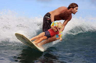 Surfista sin camisa con pantalones cortos negros surfeando una ola con un niño pequeño en brazos, quien lleva gafas de sol amarillas y flotadores de brazo con diseño colorido. Ambos disfrutan de una emocionante jornada de surf bajo un cielo claro