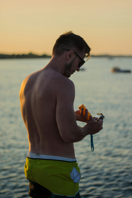 Hombre con gafas de sol y bañador verde aplicándose protector solar en el brazo, de pie en la playa al atardecer.